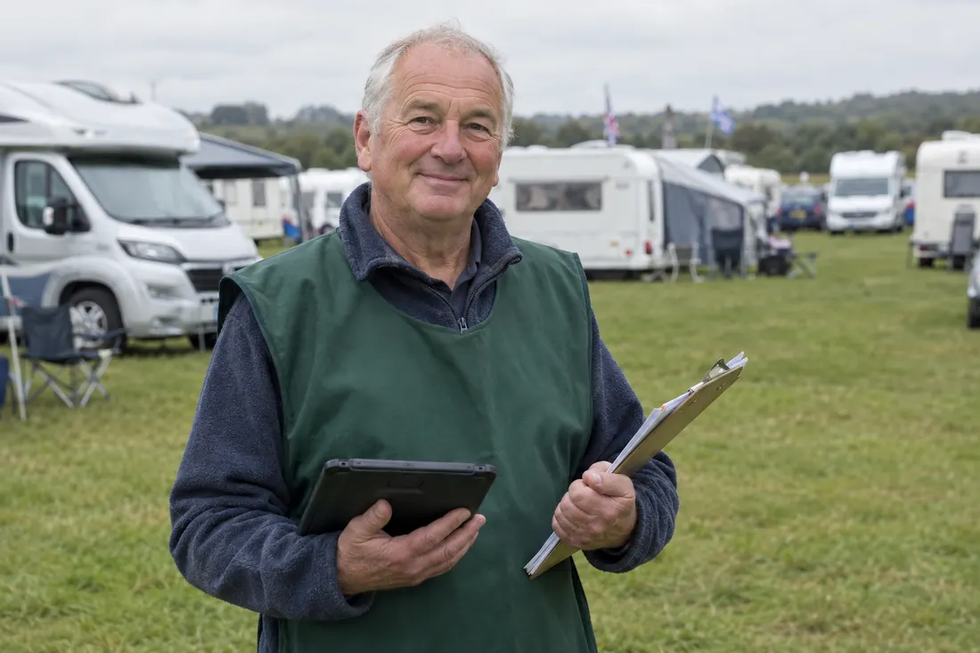 A smiling rally warden in a green tabard holding a tablet and clipboard in a field of caravans